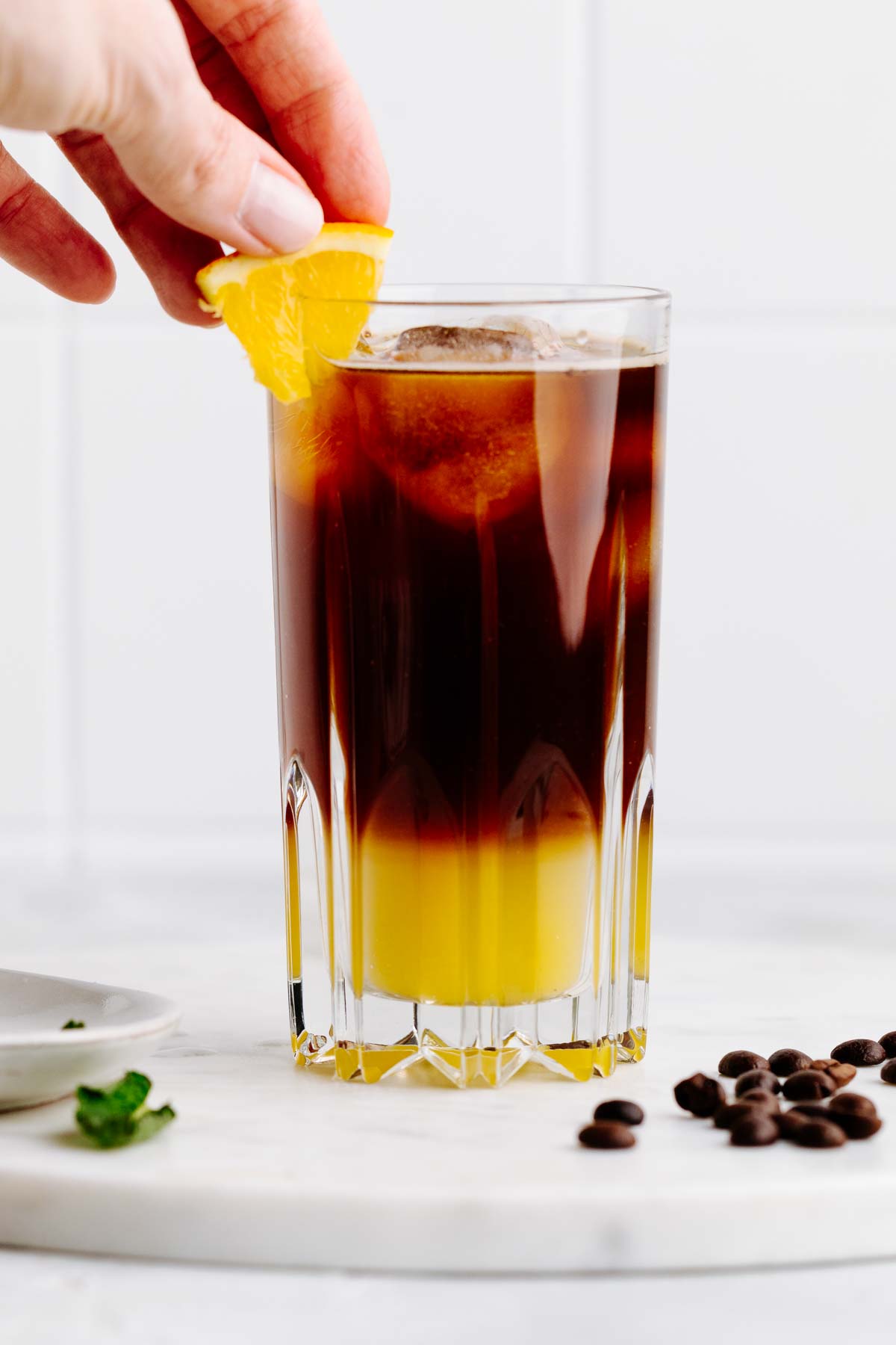 A tall glass with a two colored yellow and brown layered drink with a hand adding an orange wedge as a garnish on a white marble backdrop with coffee beans next to it.
