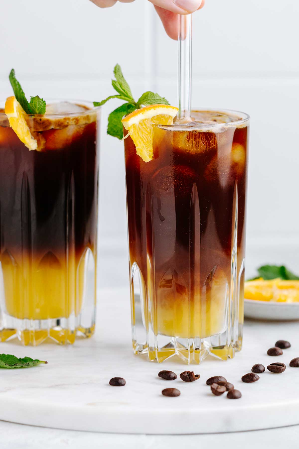 Two tall glasses with a two colored yellow and brown layered drink with a mint leave and orange wedge garnish on a white marble backdrop with coffee beans next to it and a transparant straw in one glass stirring the drink.