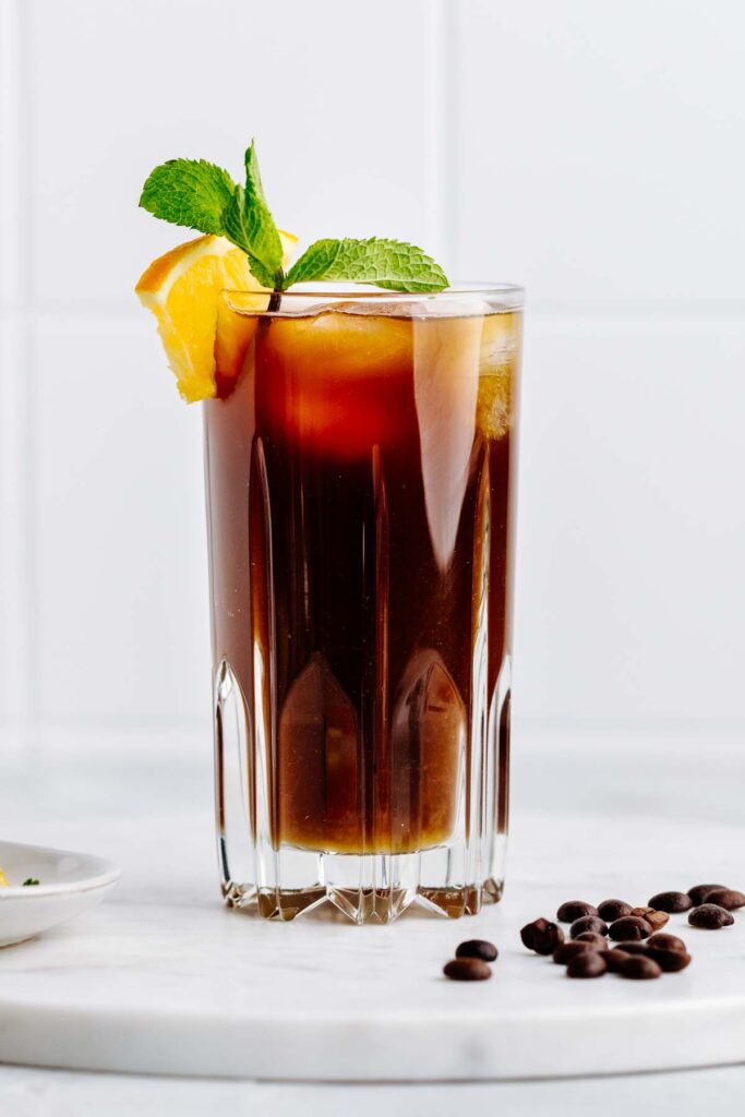 A tall glass with a brown colored drink in it with a mint leave and orange wedge garnish on a white marble backdrop with coffee beans next to it.