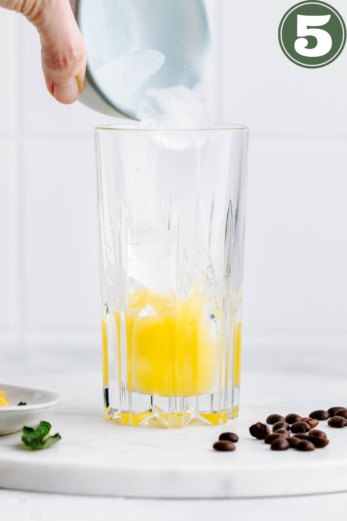 A tall glass with yellow orange juice and ice cubes in the bottom and a hand adding in more ice cubes from a small light blue bowl on a white marble backdrop with coffee beans next to it.