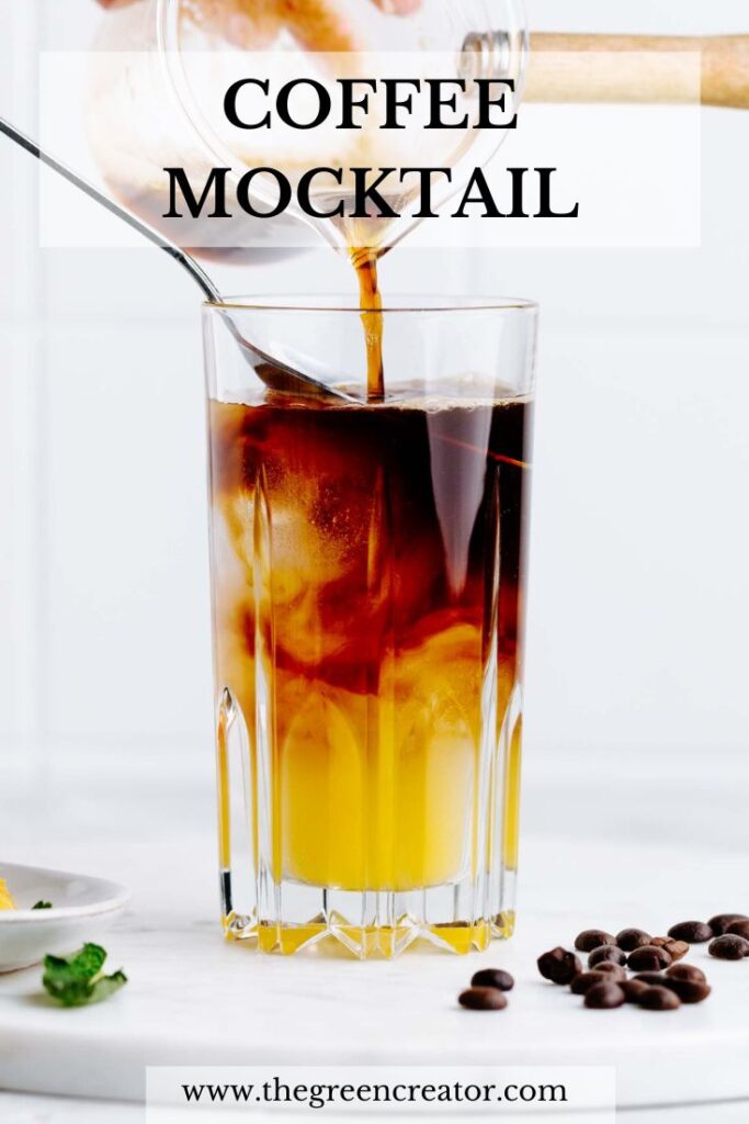 A tall glass filled halfway with two layers of yellow orange juice and brown coffee with a spoon hanging in the glass and coffee being poured over the spoon from a small transparant jug on a white marble backdrop and the text coffee mocktail written in bold on top of the photo.