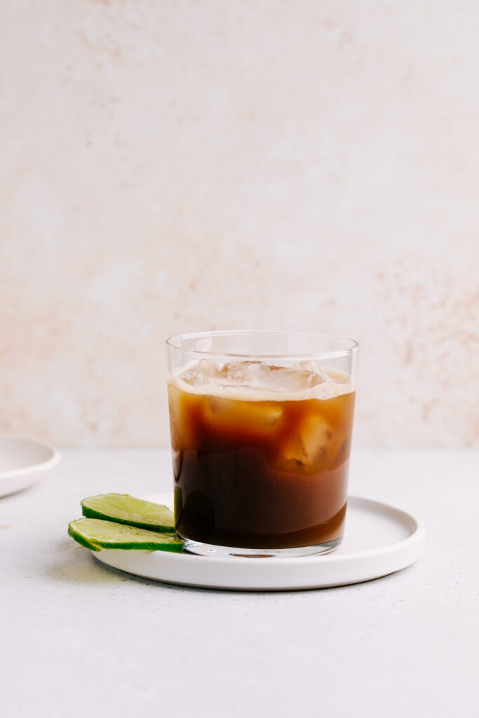 A glass with cold brew tonic on a white plate with a light brown backdrop and a few lime slices next to it