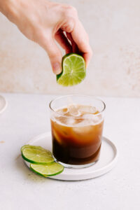 A glass with cold brew tonic on a white plate with a light brown backdrop and a few lime slices next to it with a hand squeezing in a few drops of lime juice in the drink