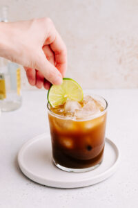A glass with cold brew tonic on a white plate with a light brown backdrop and a hand placing a lime slice in the drink