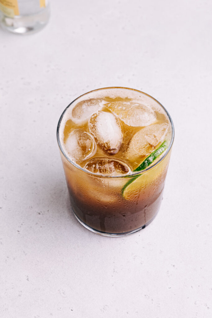 A glass with cold brew tonic with ice cubes and a slice of lime seen from slightly above on a light grey backdrop