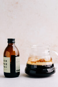 A glass coffee pot filled half with dark coffee and a dark bottle with cold brew coffee concentrate next to each other with a light brown backdrop