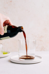 A glass on a white plate with a light brown backdrop and a hand pouring in dark cold brew coffee concentrate