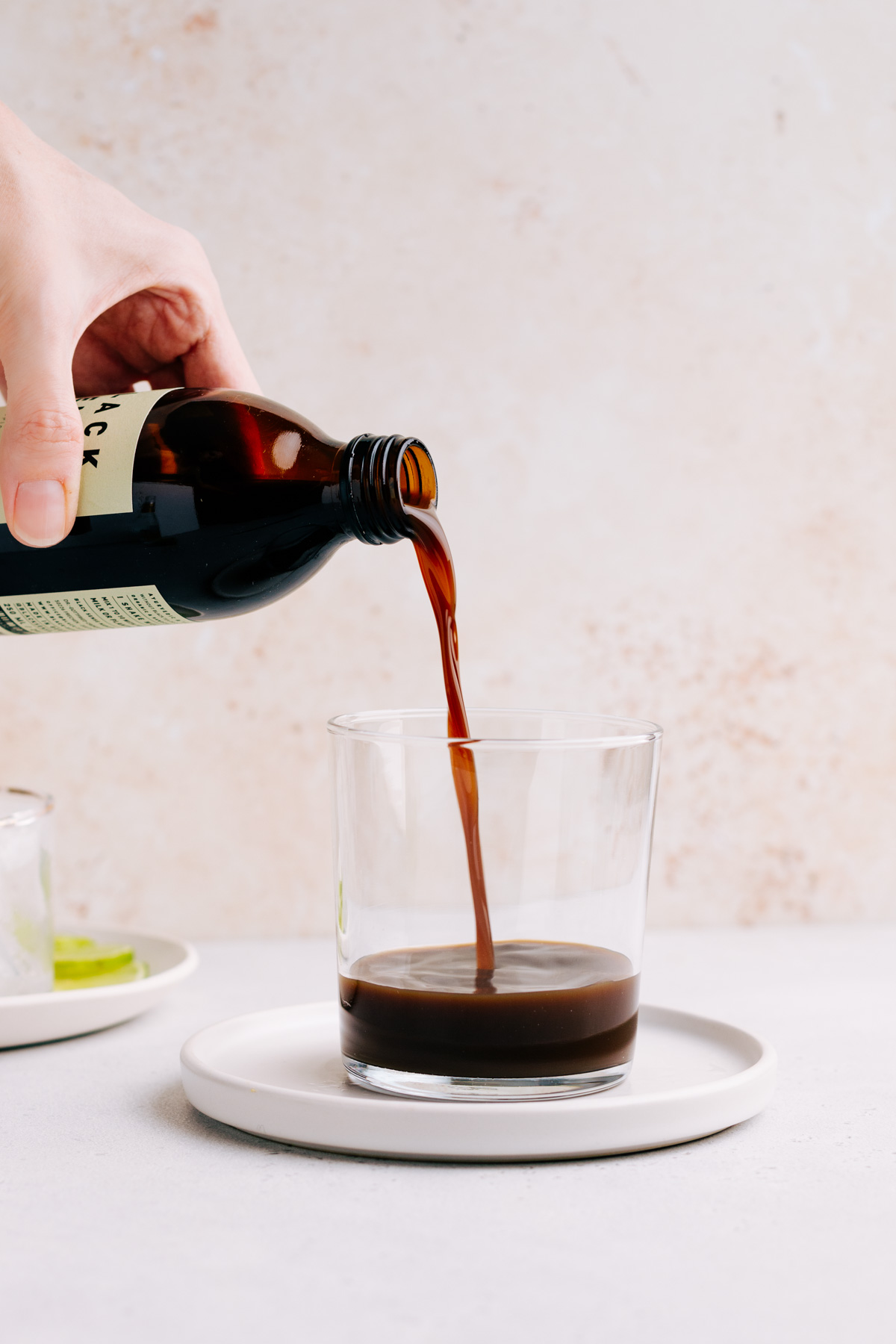 A glass with a bit of cold brew concentrate on a white plate with a light brown backdrop and a hand pouring in more cold brew coffee concentrate
