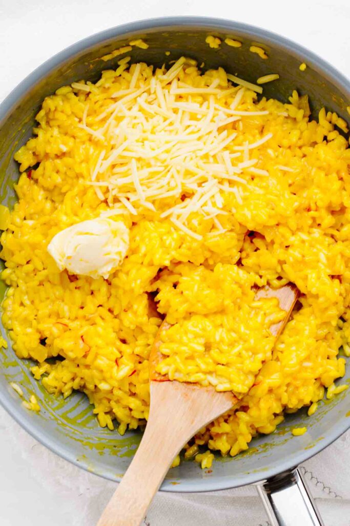 A blue colored pan with yellow saffron risotto in it and a wooden spatula in the rice with Parmesan cheese and butter on top of the rice.