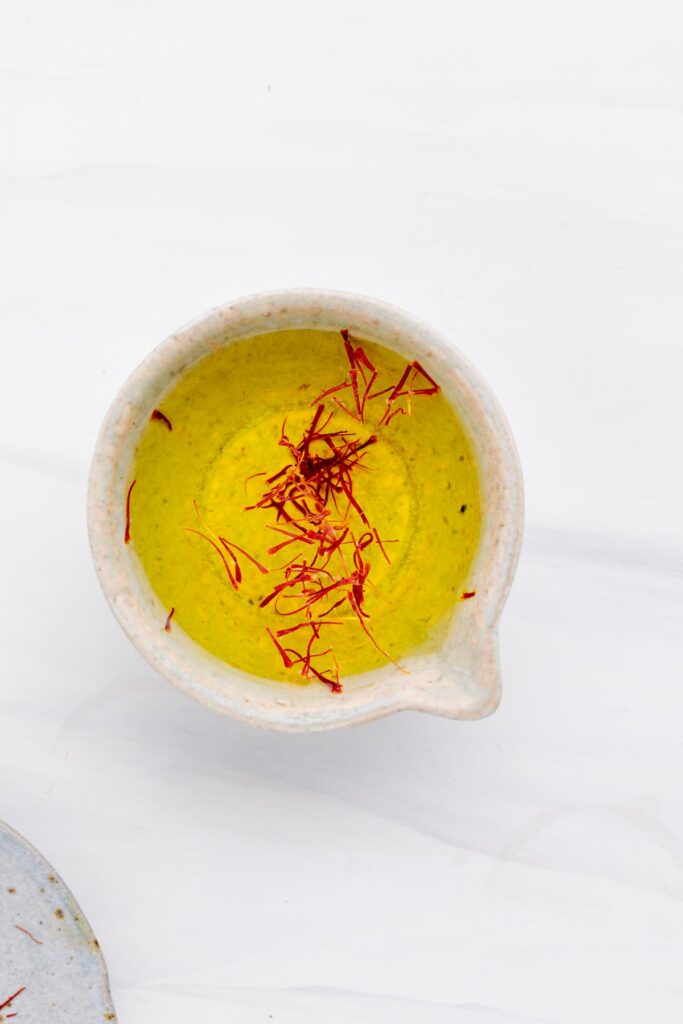 A small bowl on a white backdrop with yellow colored water in it and red saffron threads floating on top.