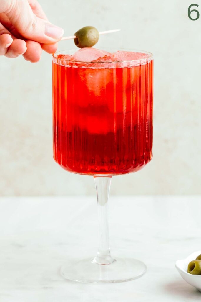 A red colored select spritz cocktail in a wine glass with ice cubes and a hand placing a green olive on a cocktail skewer as a garnish on the rim of the glass.