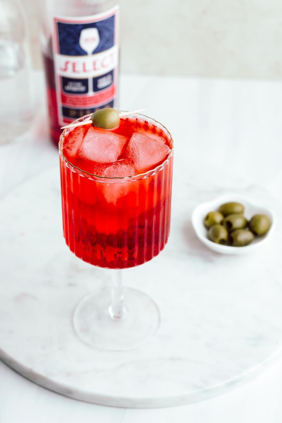 A red colored select spritz cocktail with ice cubes garnished with a green olive in a wine glass with a stem on a round white marble backdrop with olives and a bottle of select aperitivo in the background.