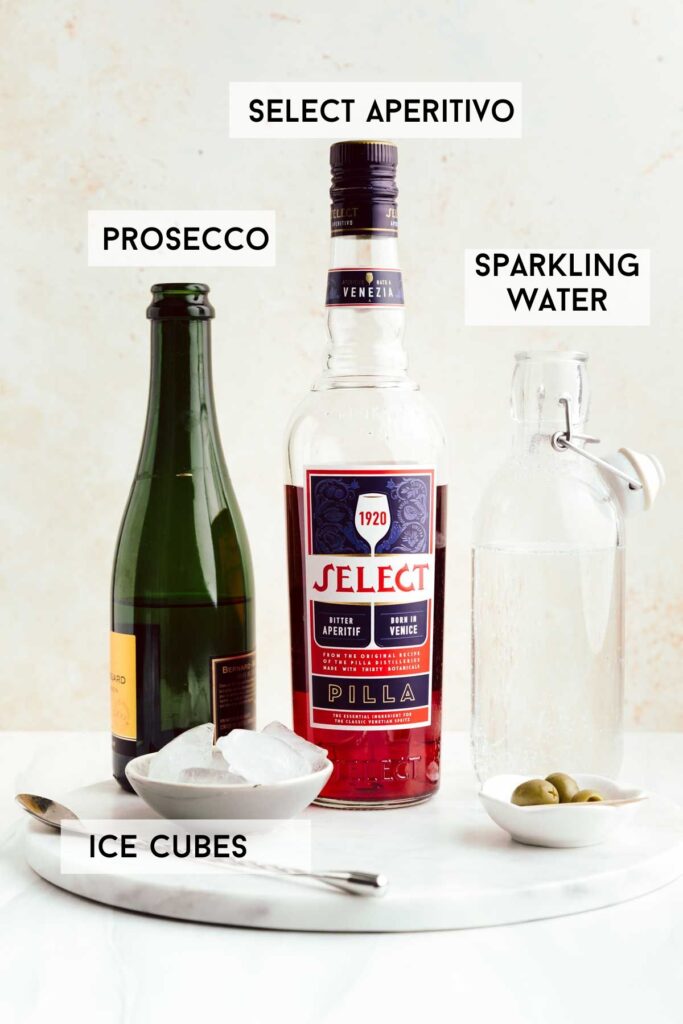 A bottle of prosecco, next to a bottle of select aperitivo and sparkling water with ice cubes and olives and a silver spoon on a round marble backdrop with the names of the ingredients next to it in bold.