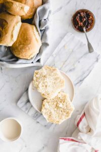 a dinner roll cut in half on a small white plate on a white marble backdrop with a small bowl with red jam, a small white mug of milk, a white napkin, a little basket with dinner rolls with a blue napkin on a white backdrop
