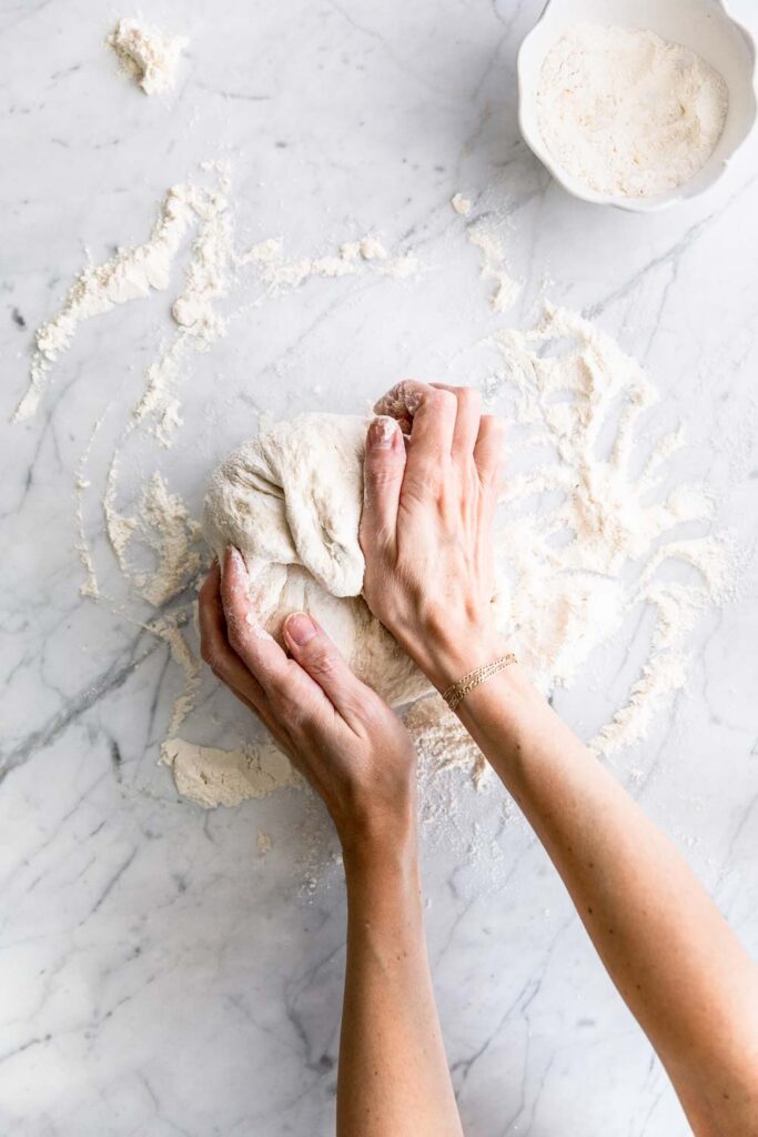 dinner rolls dough on a white backdrop with flour and a pair of hands kneading the dough