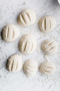 eight round dinner rolls dough on a white backdrop with flour with two cuts in each dinner roll