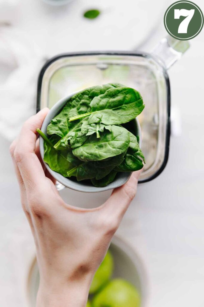A hand hovering over a blender container holding a small bowl with fresh spinach leaves in it.