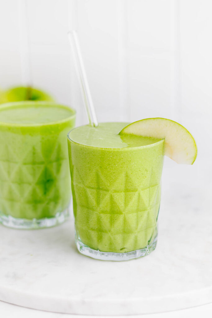 A green smoothie in a textured glass decorated with a slice of green apple and a glass straw on a white marble backdrop with green apples and another glass with green smoothie behind it.