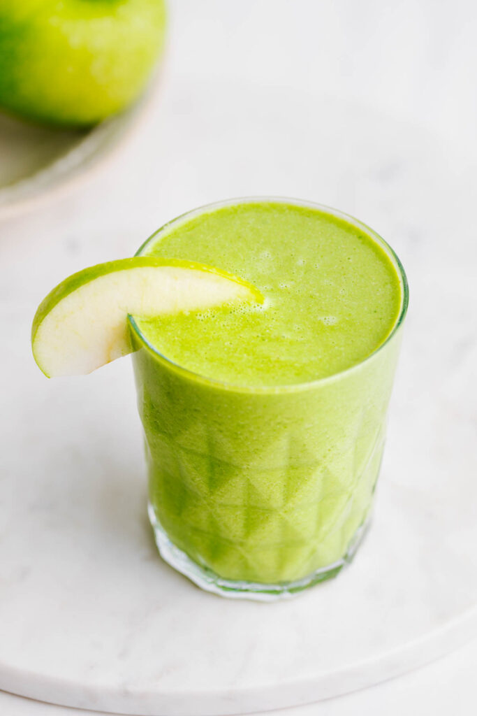 A green smoothie in a textured glass decorated with a slice of green apple on the rim of the glass on a round white marble backdrop cutting board with a green apple showing in the backdrop.