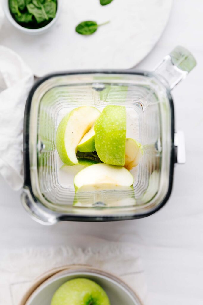 A top view of a glass blender container on a white backdrop with milk and sliced green apples in it.