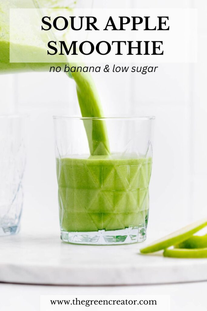 A textured glass filled half with green sour apple smoothie on a white marble backdrop with a blender container pouring in more green smoothie with the text sour apple smoothie on top of the photo.