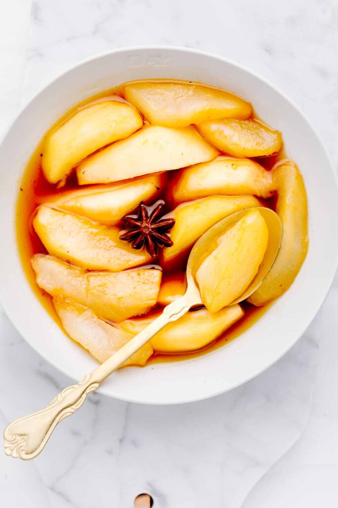 Stewed pears in an orange colored syrup in a white bowl with a star anise and a golden spoon on top of the pears.