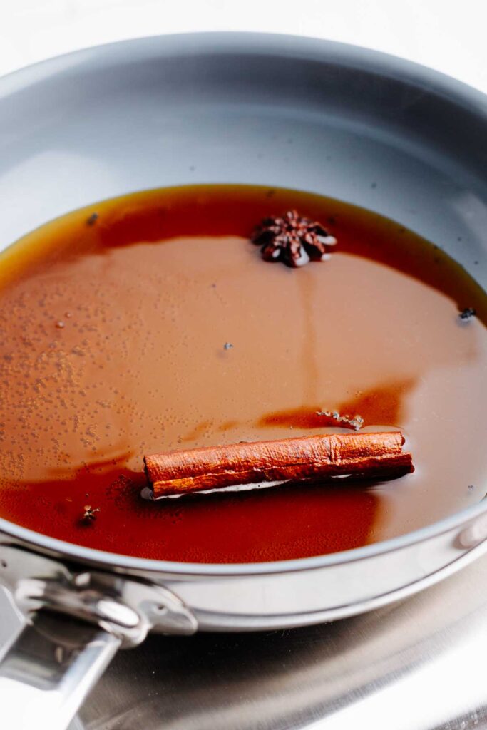 A blue pan on a small silver stove with a brown syrup, a cinnamon stick and a star anise in it.