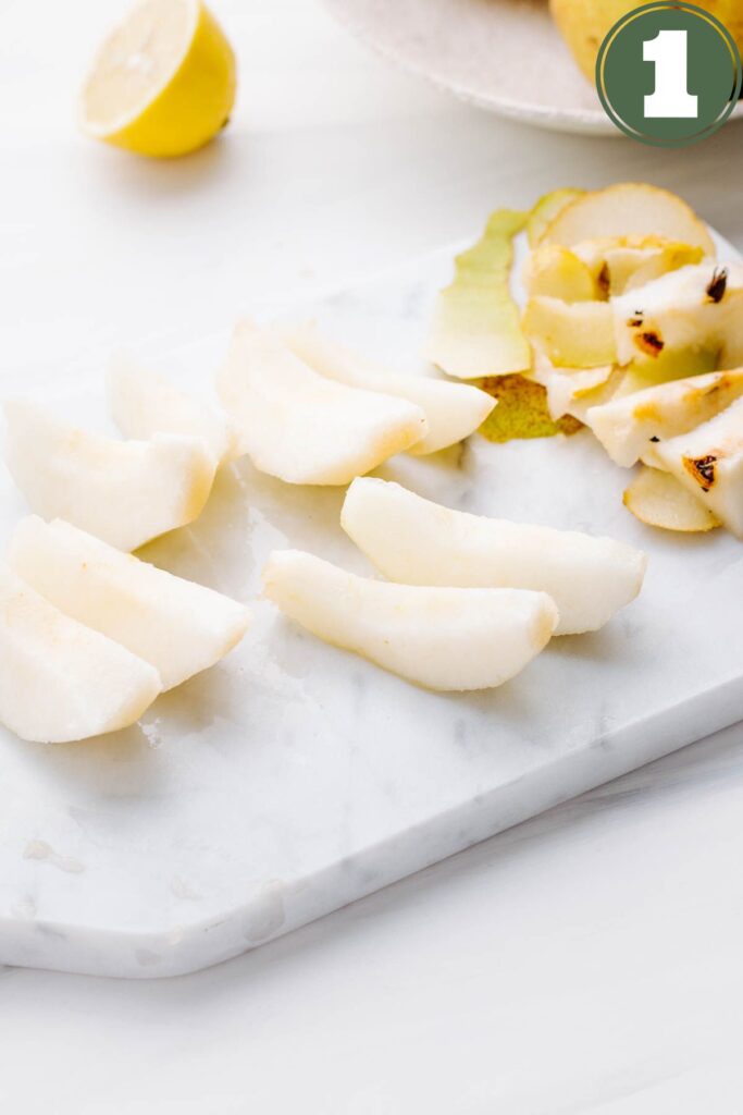 A white rectangle marble cutting board with sliced pears on it and the peel of the pears next to it with half a lemon in the backdrop.