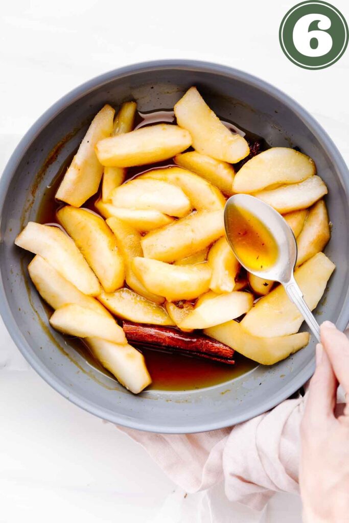 A blue pan with pears, a brown syrup and a cinnamon stick in it with a hand holding a spoon pouring the liquid over the pears.
