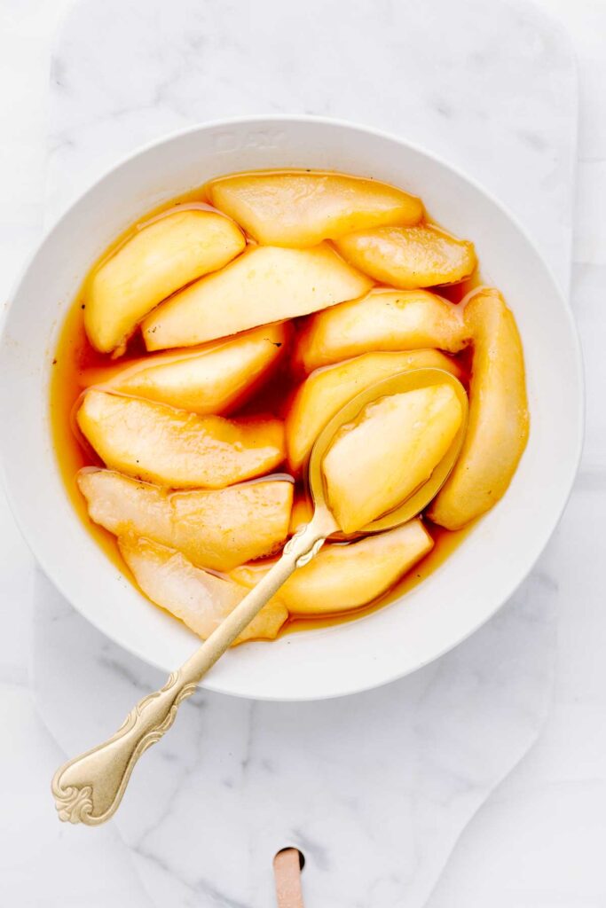 Stewed pears in an orange colored syrup in a white bowl with a gold spoon on top of the pears with one pear slice on the spoon on a white marble backdrop.