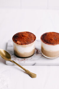 Two small jars with tiramisu overnight oats dusted with cacao powder on white marble with a gold teaspoon next to it