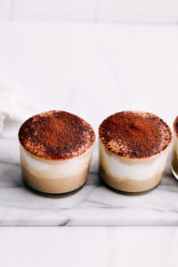 Two small jars with tiramisu overnight oats dusted with cacao powder on white marble