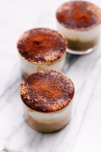 Three small jars with tiramisu overnight oats dusted with cacao powder in a row on white marble