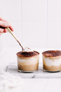 Two small jars with tiramisu overnight oats dusted with cocoa powder on white marble with a hand holding a spoon into one jar