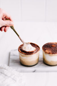 Two small jars with tiramisu overnight oats dusted with cocoa powder on white marble with a hand holding a spoon scooping up some oats and yogurt
