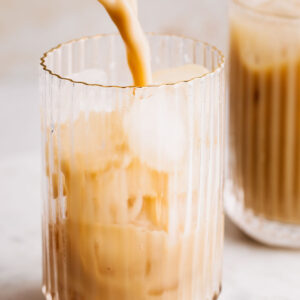 A cocktail shaker pouring in a light brown colored drink in a ribbed glass with ice cubes on a light brown backdrop with another drink in the background.