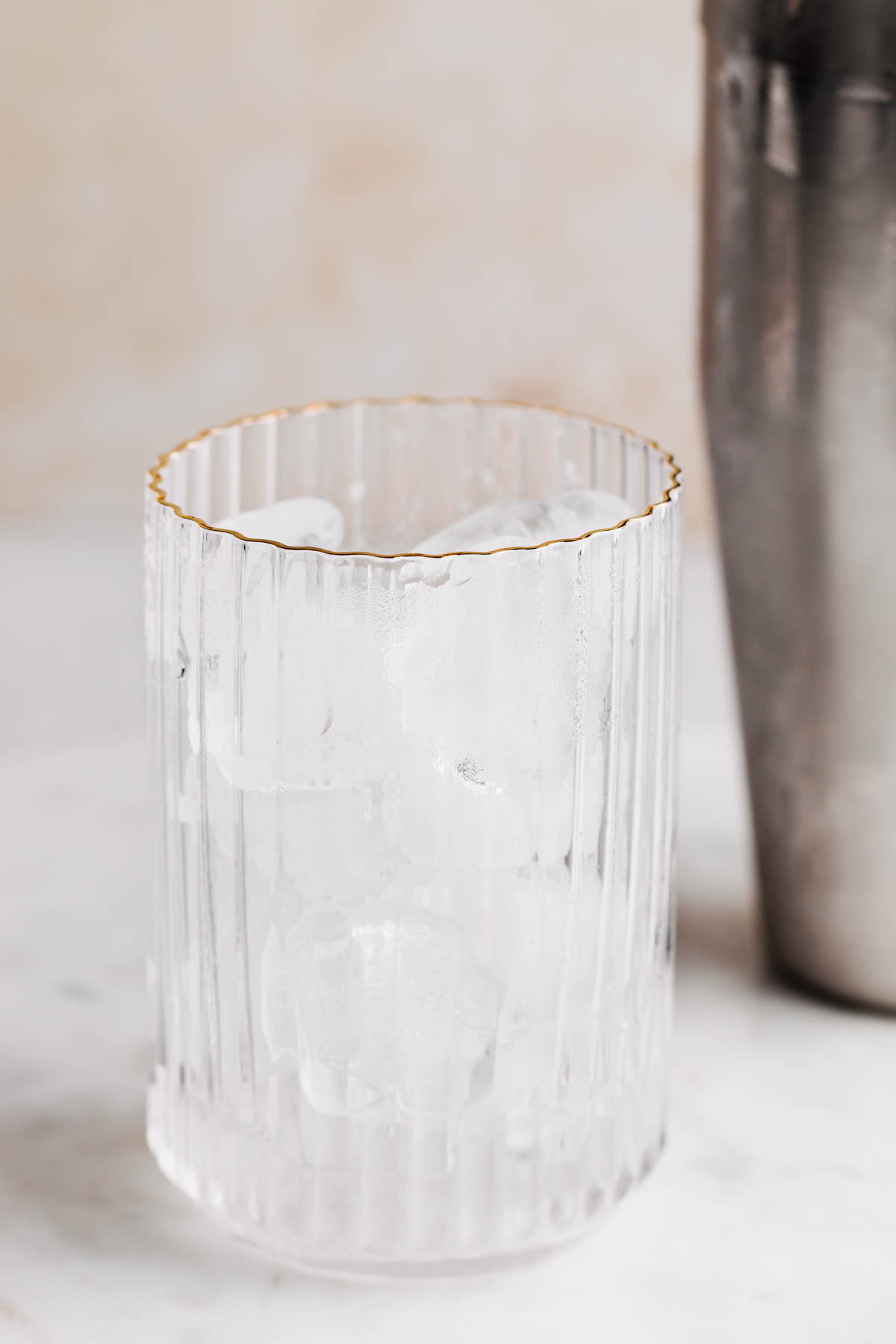 A ribbed glass with a gold rim on a light brown backdrop filled with ice cubes and a silver colored condensed cocktail shaker in the background.
