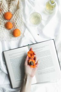 an open book with a hand on it with an open apricot in the hand on a white blanket with more apricots and drinks