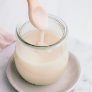 A small glass jar on a white marble backdrop with a small wooden spoon with condensed milk lifted from the glass jar with thick condensed milk dropping of the spoon in the glass.
