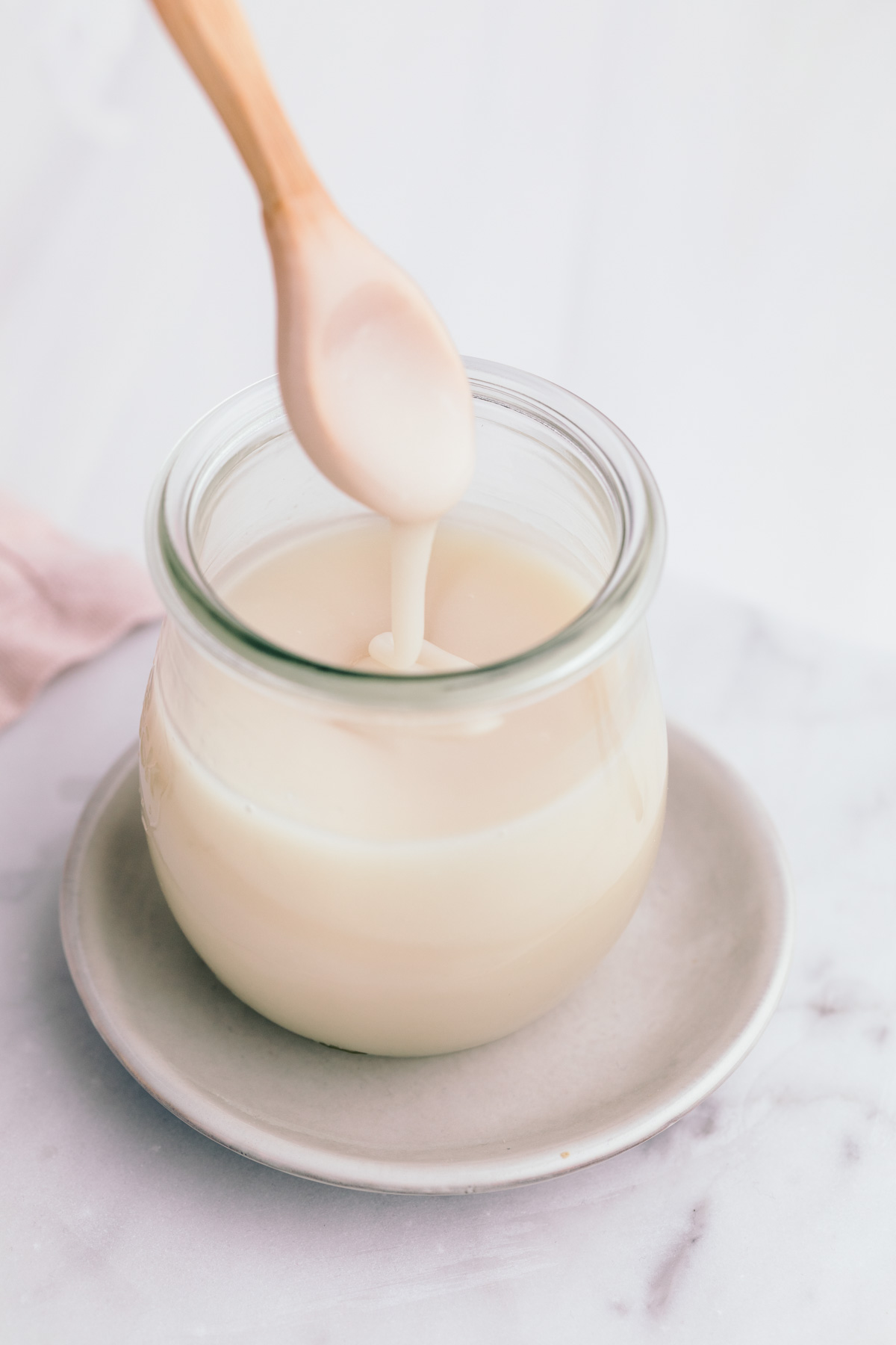 A small glass jar on a white marble backdrop with a small wooden spoon with condensed milk lifted from the glass jar with thick condensed milk dropping of the spoon in the glass.