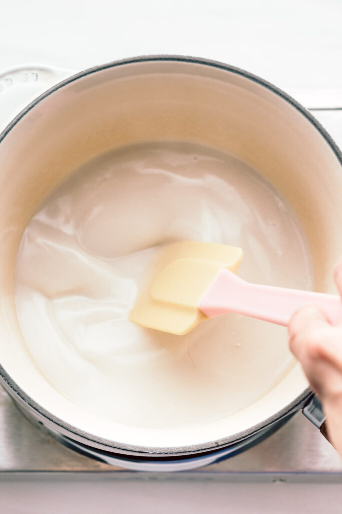 A pot on a small silver stove with dark white condensed milk in it with a hand stirring the milk in the pot with a spatula.