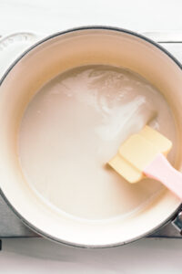 A pot on a silver stove with thick light yellow condensed milk in it with a spatula in the pot stirring the milk.