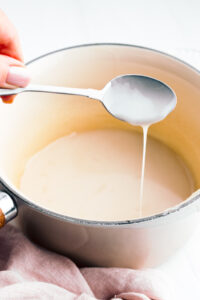 A pot with light yellow condensed milk in it next to a pink napkin and a hand holding a silver spoon above the pot with dripping thick milk.