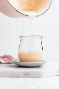 A pan pouring in thick condensed milk in a glass jar on a small plate on a white marble cutting board with a white backdrop.