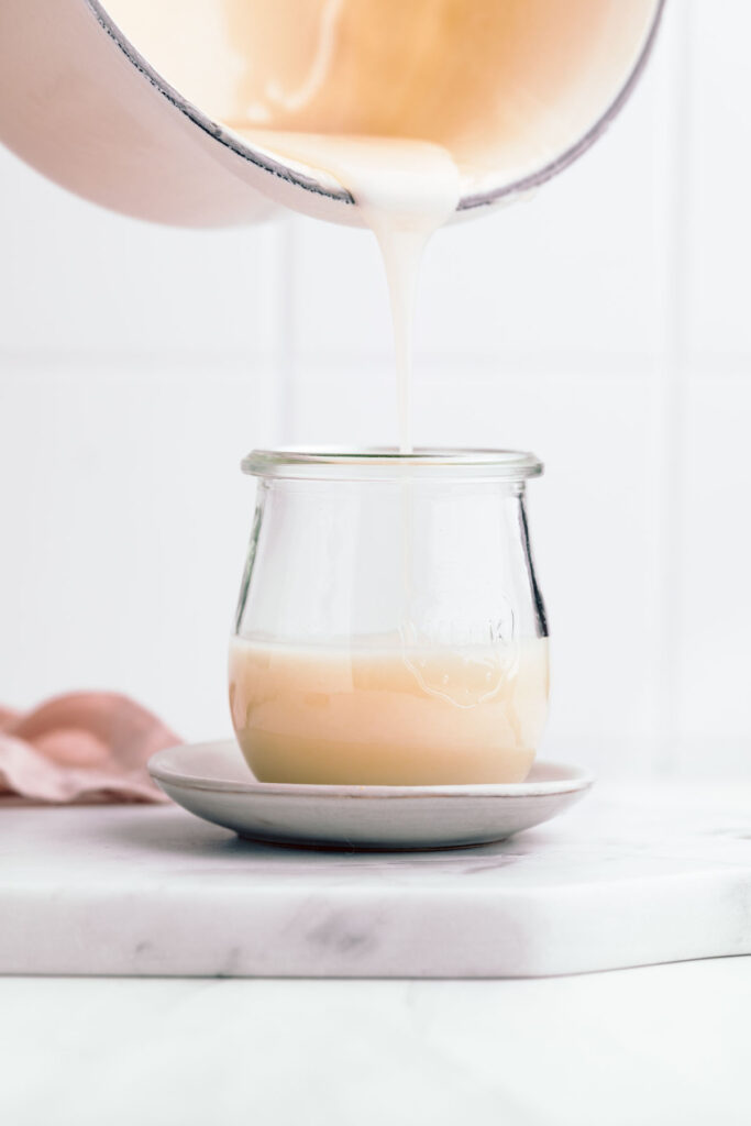 A pan pouring in thick condensed milk in a glass jar on a small plate on a white marble cutting board with a white backdrop.