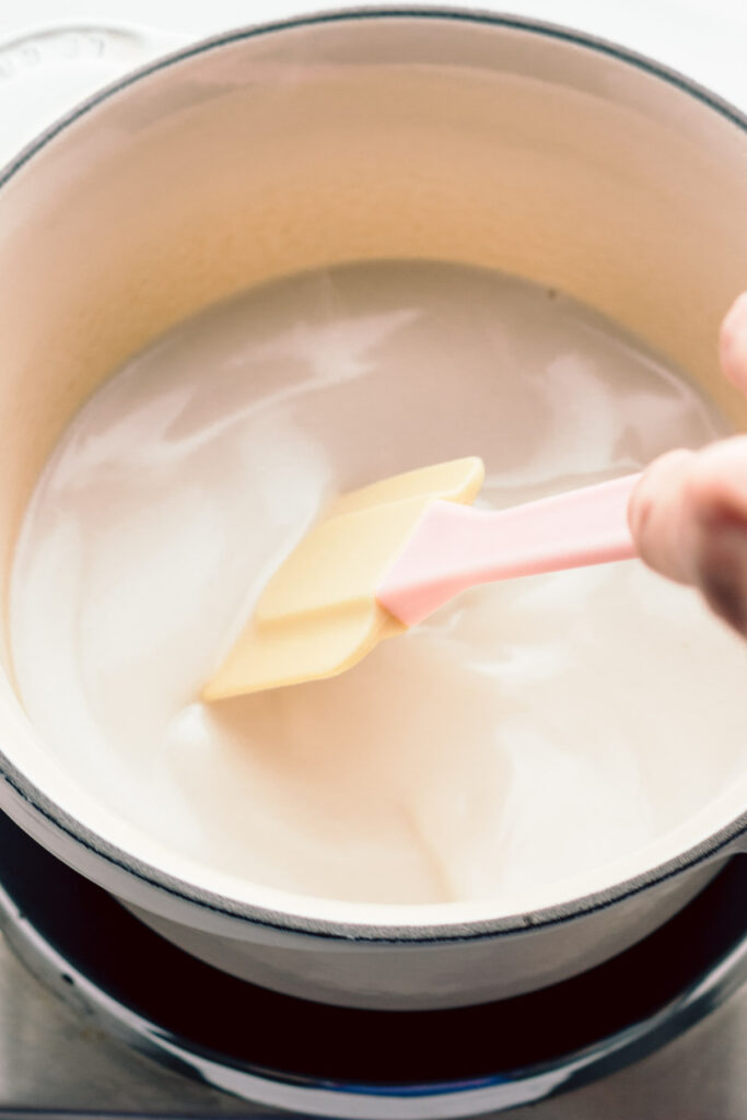 A pot with dark white condensed milk in it and a spatula with a hand stirring the milk in the pot.