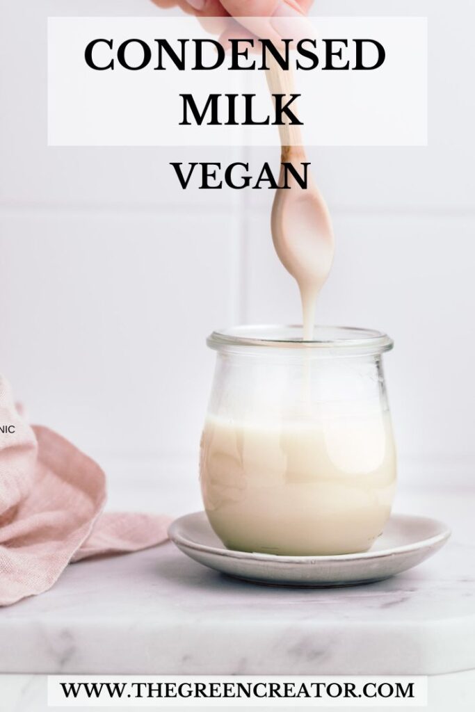 A small glass jar on a white marble backdrop with a small wooden spoon above the glass jar with thick condensed milk dripping in the glass and the text condensed milk vegan written on top of the photo.