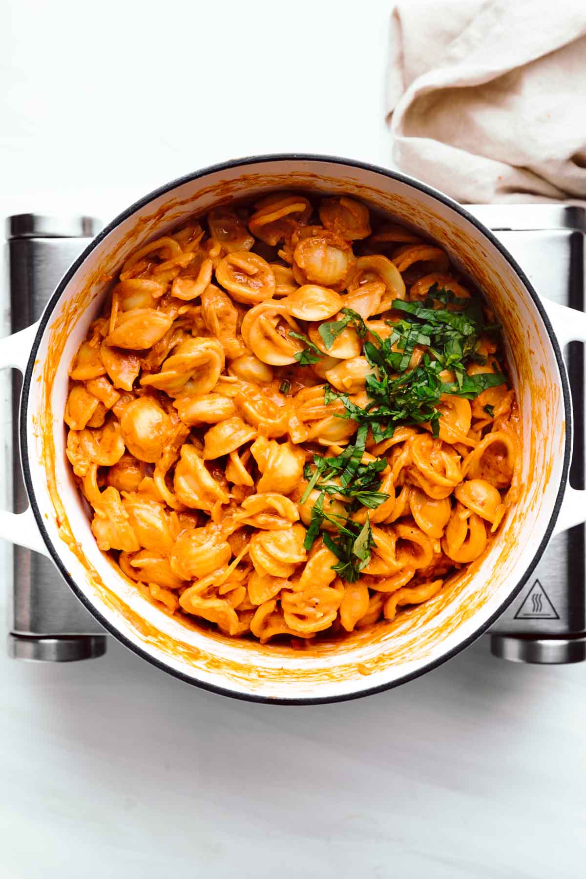 A large white pot with orange colored shell pasta in the pot sprinkled with chopped green basil on a little stove with a napkin next to it.