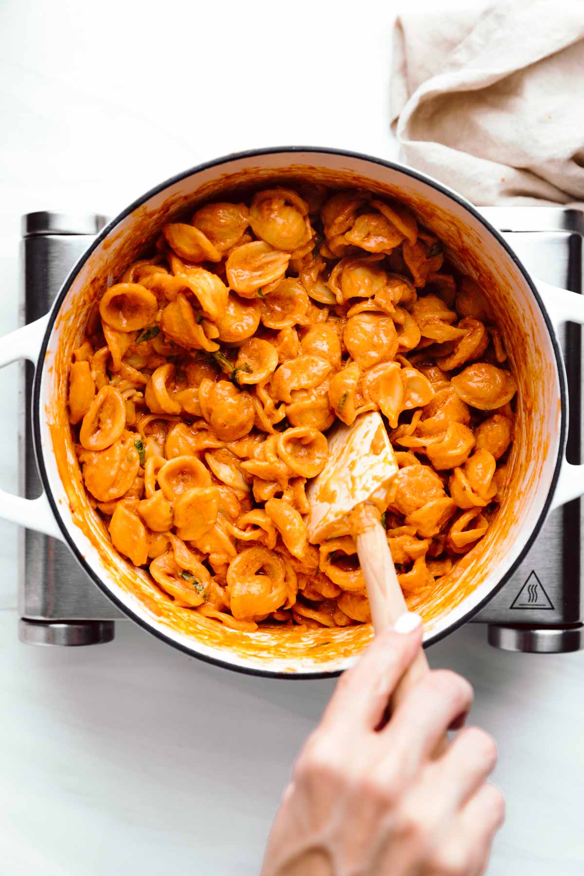 A large white pot with orange colored shell pasta and a hand with a spatula in the pasta stirring the pasta on a little stove with a napkin in the top corner.