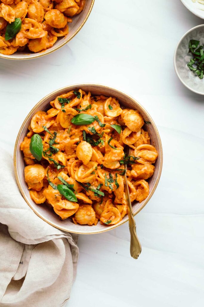 Orange colored shell pasta in a bowl with a golden utensil topped with chopped basil and small basil leaves on a white backdrop next to a beige colored napkin and a part of another bowl with pasta visible in the up left corner.
